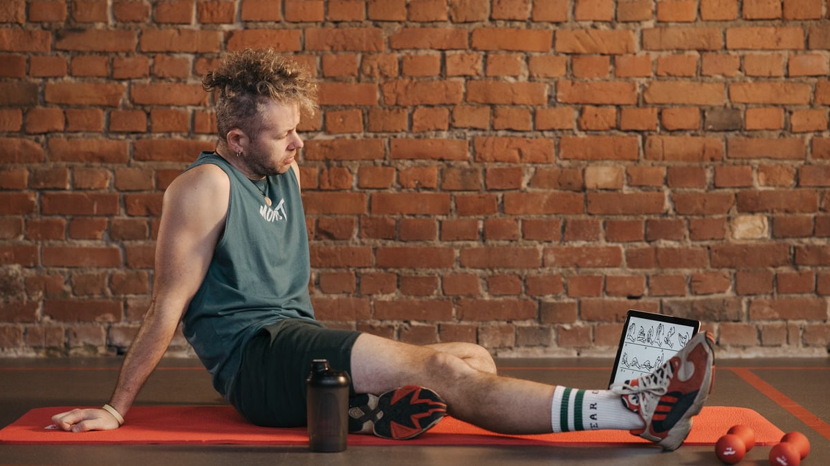 Athletic man sitting and resting on a mat in an urban gym, with a tablet and weights visible.